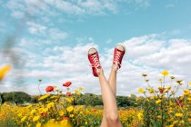 Coltivare femmina irriconoscibile in calzature luminose sdraiato con le gambe incrociate tra margherite in fiore sotto cielo blu nuvoloso in campagna — Foto stock