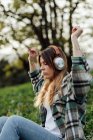 Side view of female listening to music from headphones while having fun sitting with eyes closed on summer meadow against mounts — Fotografia de Stock