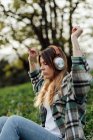 Side view of female listening to music from headphones while having fun sitting with eyes closed on summer meadow against mounts - foto de stock