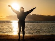Back view silhouette of traveler with arms raised and peace gesture enjoying freedom while standing on seashore at sunset time — Foto stock