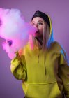 Уверенная женщина-хипстер в толстовке курит сигарету в студии на розовом фоне и отворачивается — стоковое фото