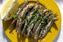 Top view plate of appetizing grilled anchovies served on table with piece of lemon and fresh herbs in restaurant — Foto stock