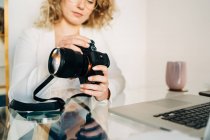Cropped serious young woman with curly blond hair in stylish outfit and eyeglasses inserting SD card into photo camera after transferring files to laptop at home — Stock Photo