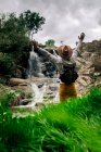 Voltar ver anônimo masculino mochileiro em chapéu apreciando vista de cascata streaming de rocha áspera na natureza verdejante — Fotografia de Stock