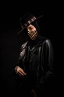 Модная исламская женщина в кожаной куртке, хиджабе и модной шляпе, стоящая перед камерой в темной студии во время вспышки коронавируса — стоковое фото