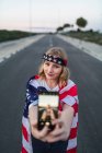 Патриотичная американка, завернутая в национальный флаг США, делает селфи на мобильном телефоне, стоя на дороге — стоковое фото