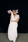 Charming young female model in bohemian elegant white lace gown and floral wreath standing against black background with neon inscription — Photo de stock