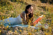 Мечтательная очаровательная брюнетка в белом платье, лежащая на полевом лугу и читающая книгу при солнечном свете — стоковое фото