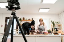Неузнаваемая женщина фотографирует шоколадные кексы на цифровой камере против блогера, разговаривающего во время приготовления пищи на кухне — стоковое фото