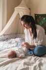 Усміхнена мама взаємодіє з невпізнаваною маленькою дитиною на збитому ліжку вдома в денний час — стокове фото