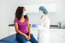 Fröhliche Ärztin in Schutzuniform, Latexhandschuhen und Gesichtsmaske im Gespräch mit einer afroamerikanischen Patientin vor der Impfung in der Klinik während des Coronavirus-Ausbruchs — Stockfoto