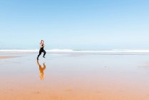 Vista laterale del jogger femminile attivo con tatuaggi che corrono sulla riva dell'oceano mentre riflettono in acqua durante l'allenamento — Foto stock