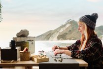 Seitenansicht von zufriedenen Reisenden, die handgefertigte Accessoires kreieren, während sie am Holztisch in einem geparkten LKW am Meer sitzen — Stockfoto