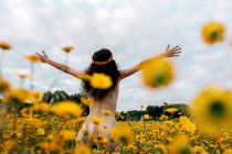 Back view of anonymous naked brunette in flower wreath enjoying meadow with blossoming daisies under cloudy sky in summer — Stock Photo