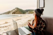 High angle of female traveling freelancer sitting in parked truck with smartphone and laptop and working on remote project while looking away — Stock Photo