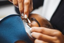 Crop unrecognizable beauty technician with cotton bud preparing ethnic woman with patch for eyelash extension procedure in salon — Photo de stock