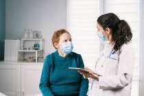 Sanitäterinnen in Uniform mit Tablette sprechen mit Seniorin in steriler Maske auf Konsultation, während sie sich während der Coronavirus-Pandemie anschauen — Stockfoto