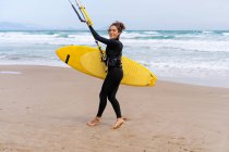Sportswoman en combinaison avec cerf-volant gonflable se promenant sur le rivage sablonneux tout en regardant la caméra contre l'océan orageux — Photo de stock