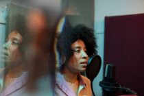 Cantante femenina negra interpretando canción contra micrófono con filtro pop mientras está de pie y mirando hacia adelante en el estudio de sonido - foto de stock
