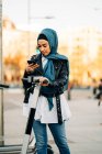 Мусульманская женщина в традиционном платке с помощью приложения на смартфоне и сканирующего дисплея для разблокировки современного электрического скутера, припаркованного на городской улице — стоковое фото