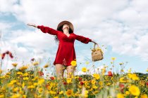 Снизу счастливая женщина в красном сарафане, шляпе и сумочке стоит с закрытыми глазами на цветущем поле с желтыми и красными цветами с протянутыми руками, наслаждаясь теплым весенним днем — стоковое фото