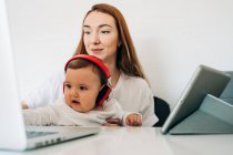 Positiva joven madre y lindo bebé atento en los auriculares viendo dibujos animados en el ordenador portátil mientras están sentados en el escritorio juntos - foto de stock