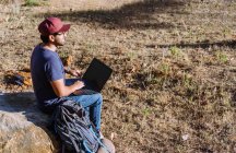 Seitenansicht Ganzkörper-Nomade sitzt auf Stein in der Nähe von Habseligkeiten und nutzt Netbook, während er aus der Ferne arbeitet — Stockfoto