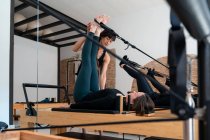 Flexible female stretching legs with help of personal instructor while doing exercises on pilates reformer — Stock Photo