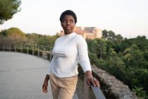 Contenuto giovane donna afroamericana in abiti casual in piedi vicino alla recinzione nel lussureggiante parco cittadino durante la giornata estiva e guardando altrove — Foto stock