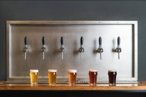 Verschiedene Biersorten mit Schaumstoff in Glaskrügen gegen Reihe von Zapfhähnen in Bar auf grauem Hintergrund — Stockfoto