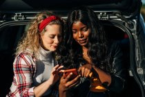 Joyeuses amies multiraciales assises dans le coffre de la voiture et naviguant sur les médias sociaux sur le téléphone mobile ensemble — Photo de stock