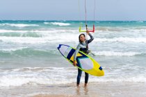 Female athlete in wetsuit with control bar looking at camera on sandy shore against foamy ocean after practicing kiteboarding — Stock Photo