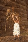 Веселый очаровательный ребенок в комбинезоне играет с сеном возле соломенных тюков в сельской местности — стоковое фото