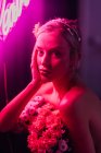 Attraktive junge barbusige Frau in weißen Hosen mit einem Strauß frischer bunter Blumen sitzt auf einem Hocker vor heller Neonbeschriftung im dunklen Studio — Stockfoto