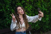 Jeune femme ludique avec de longs cheveux bouclés montrant la langue et le signe v tout en prenant selfie sur smartphone dans un parc luxuriant — Photo de stock