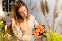 Florista femenina joven concentrada en delantal y anteojos que arregla flores amarillas fragantes en jarrón mientras trabaja en una tienda de flores - foto de stock