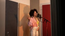 Black female singer performing song against microphone with pop filter while standing with hand on hip and looking forward in sound studio — Stock Photo