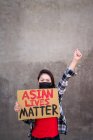 Mujer étnica en máscara y con cartel de cartón con inscripción Asian Lives Matter protestando con el brazo levantado en la calle de la ciudad y mirando a la cámara - foto de stock