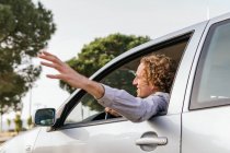 Side view of cheerful young haired male keeping hand out of car window while enjoying summer journey in nature — Foto stock