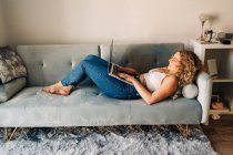 Вид на молодую самозанятую женщину в повседневной одежде и очках лежащую на удобном диване и работающую дистанционно на ноутбуке — стоковое фото