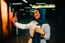 Этническая женщина в хиджабе стоит на платформе на вокзале и делает селфи на мобильном телефоне, ожидая поезда — стоковое фото