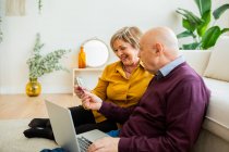 Munteres älteres Paar, das beim Online-Einkauf zu Hause per Laptop mit Plastikkarte bezahlt — Stockfoto