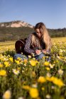 Молодая хипстерша, сидящая на лугу в сельской местности, пишет песни на блокноте и играет на гитаре во время летнего солнечного света. — стоковое фото