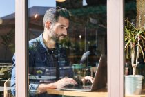 Through glass view of adult bearded ethnic male remote employee working on netbook at table — Stock Photo