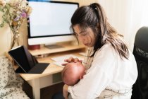 Erwachsene Mutter sitzt am Schreibtisch, arbeitet am Desktop-Computer und macht sich Notizen im Notizbuch, während sie ihr kleines Kind tagsüber am Tisch hält — Stockfoto