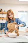 Conteúdo jovem fêmea em avental chicoteando creme doce enquanto cozinha à mesa em casa leve — Fotografia de Stock