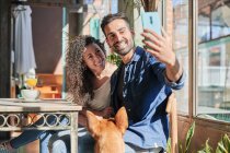 Homme ethnique souriant avec petite amie prenant autoportrait sur téléphone portable à table avec boissons contre chien au restaurant — Photo de stock