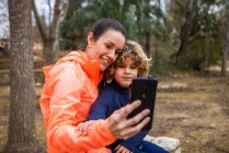 Cheerful mom in sports clothes embracing charming boy while taking self portrait on cellphone in park - foto de stock