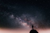 Baixo ângulo de silhueta de turista anônimo em pé no penhasco contra o céu estrelado brilhante à noite — Fotografia de Stock