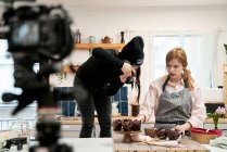 Mujer irreconocible tomando fotos de magdalenas de chocolate en cámara digital contra blogger hablando durante el proceso de cocción en la cocina - foto de stock